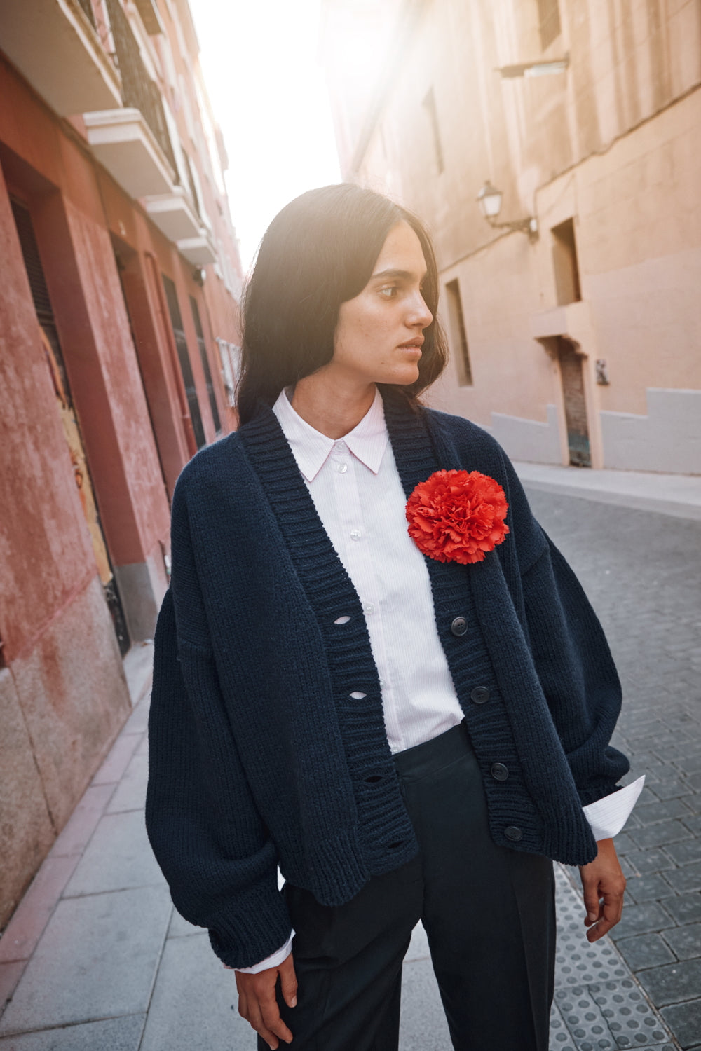 Woman wearing a navy cardigan over a white shirt with a red flower accessory, standing on a street.