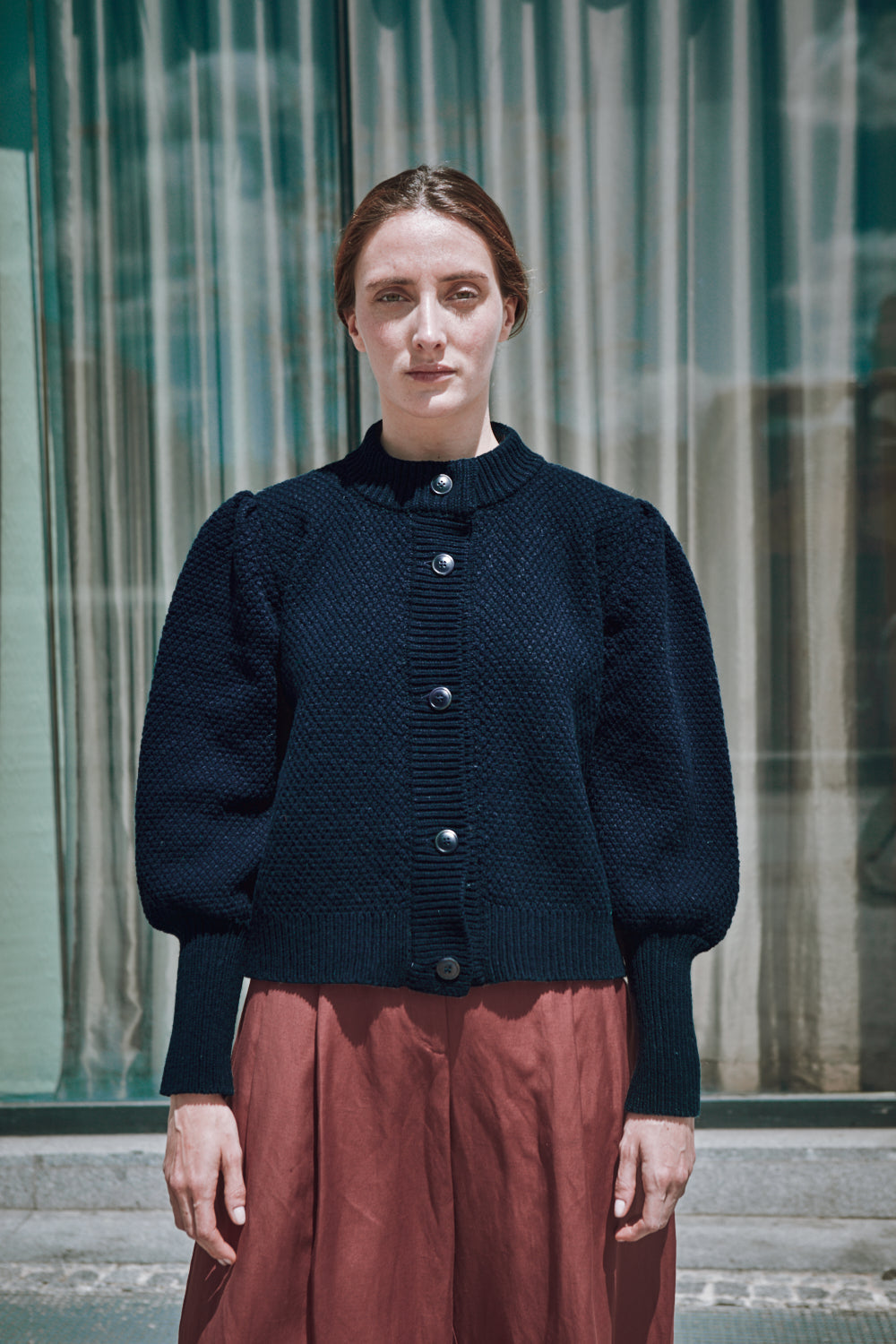 Woman wearing a dark blue  L'ENVERS cardigan and red skirt standing in front of a glass building.