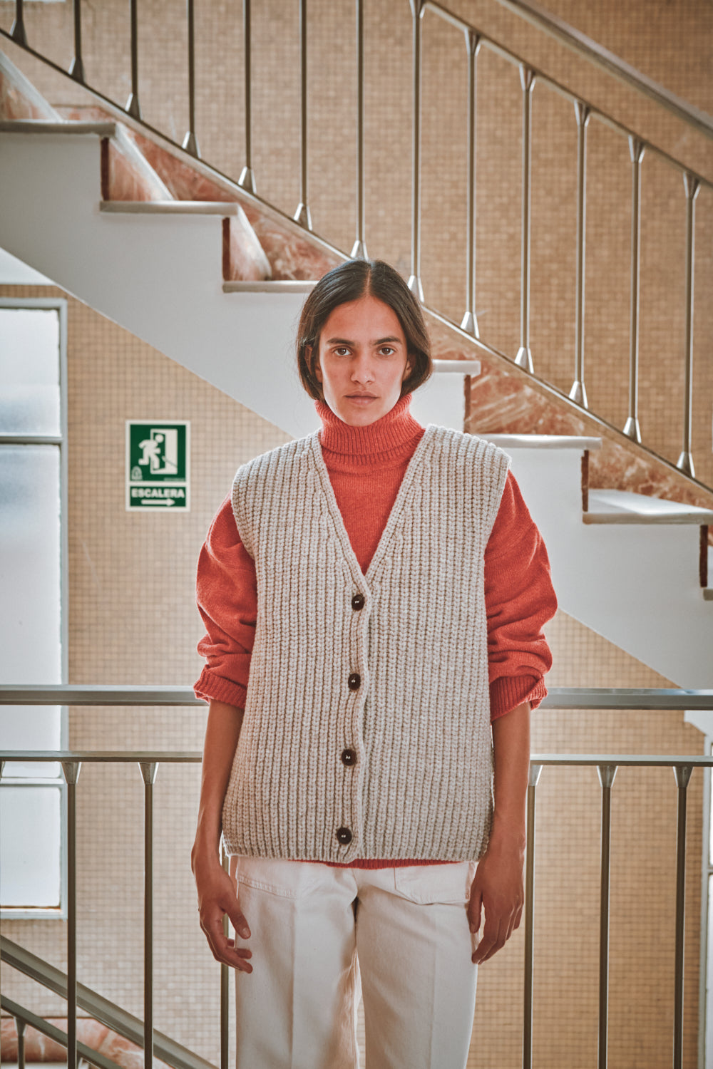 Person wearing a patterned vest over a red sweater in an indoor setting with stairs.