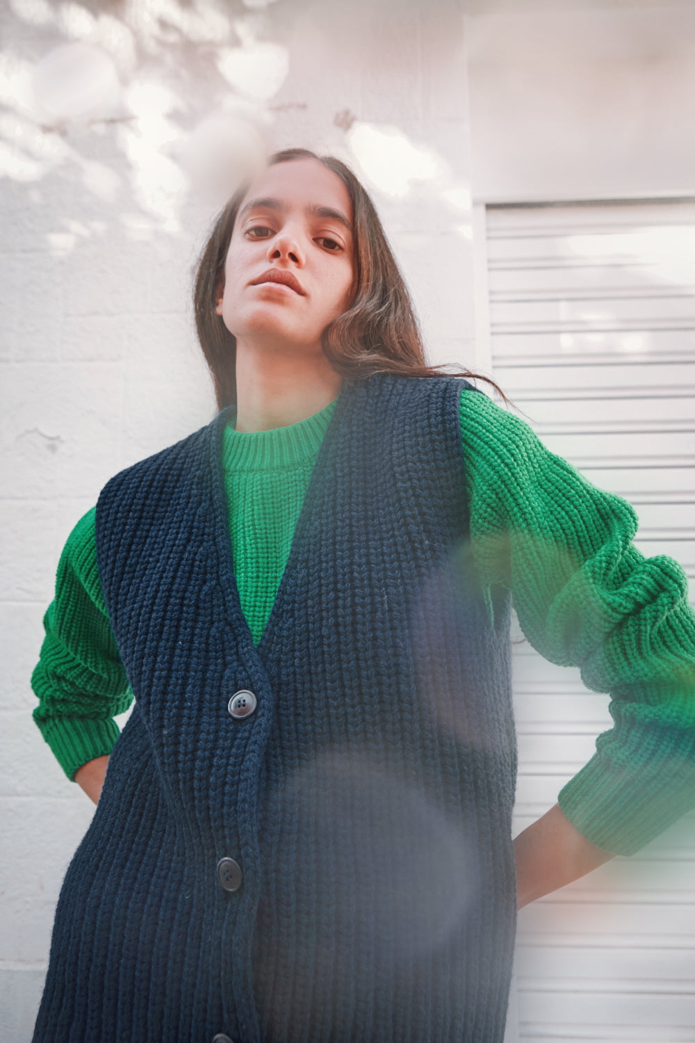 Person wearing a green sweater and dark blue vest against a white brick wall.