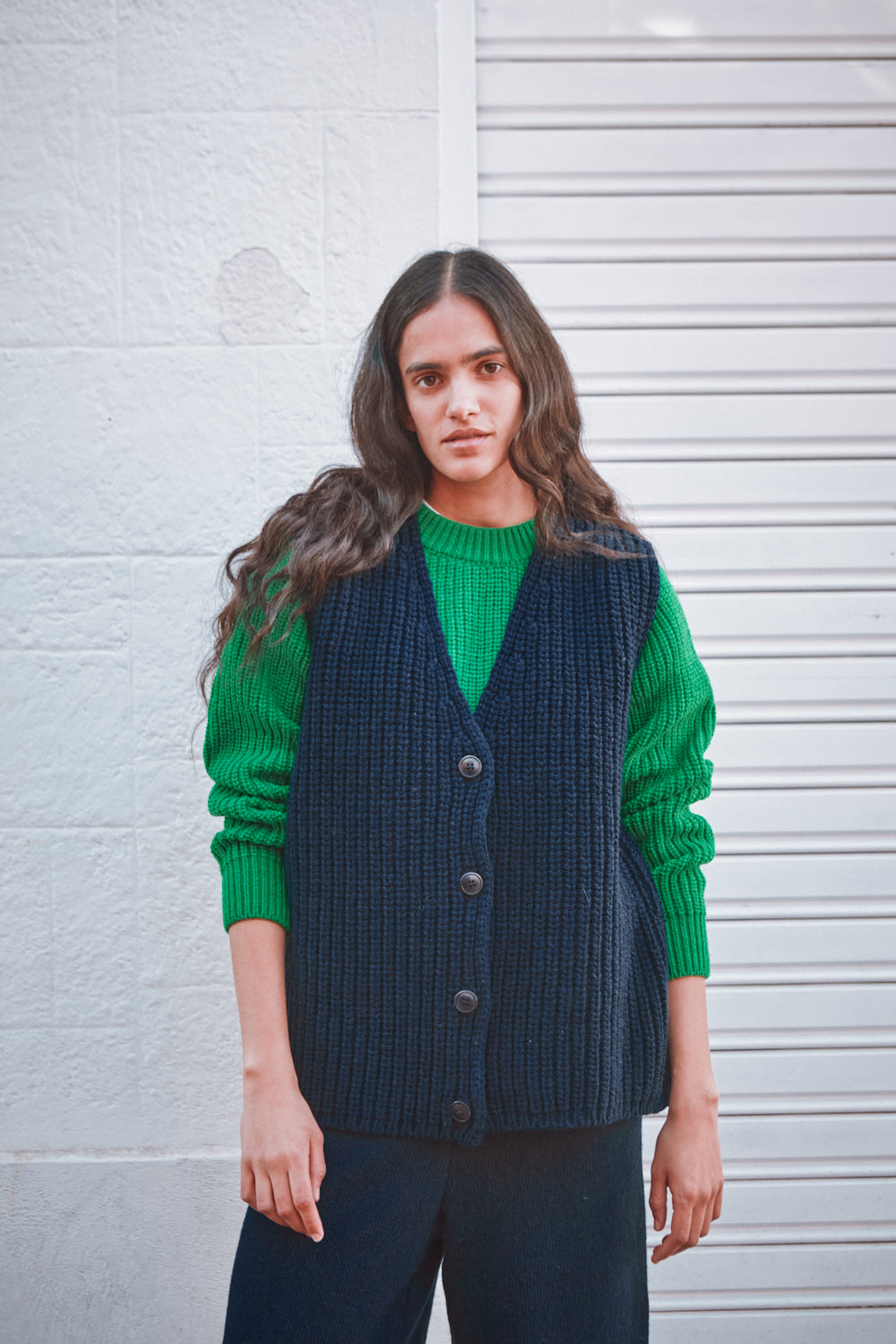 Woman wearing a green sweater and navy vest against a white wall.
